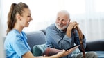 A smiling woman in a blue shirt shows a book to an elderly man with a cane on the couch.