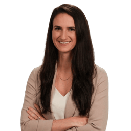 Smiling woman with long brown hair in a light blazer and white top, arms crossed, on a plain background.