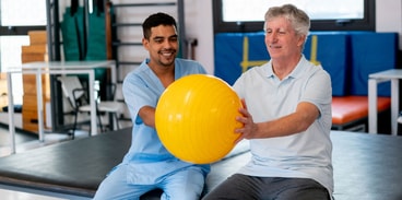 A young therapist helps an older man with a yellow exercise ball in a gym, using medbridge resources.