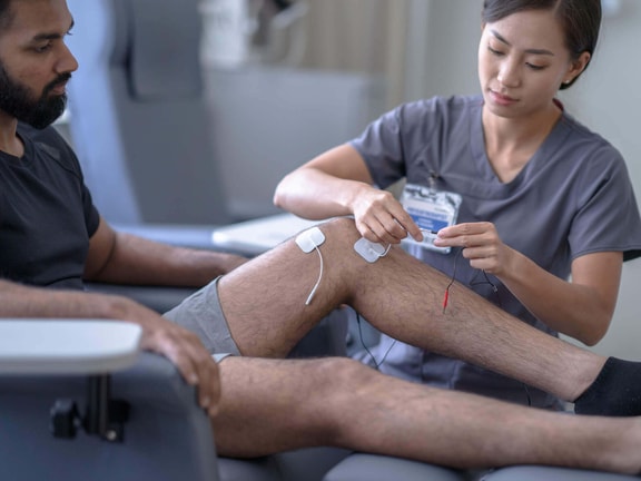 A clinician applying neuromuscular electrical stimulation (NMES) electrodes to a patient's quadriceps to address arthrogenic muscle inhibition (AMI) after knee surgery.