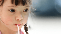 A young girl with braids and a colorful headband sips a drink through a straw while looking to the side against a blurred background.