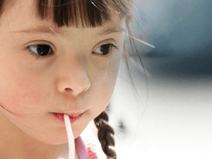A young girl with braids and a colorful headband sips a drink through a straw while looking to the side against a blurred background.