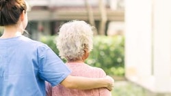 A caregiver in blue scrubs walks with an elderly person outside on a sunny day.
