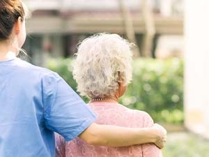 A caregiver in blue scrubs walks with an elderly person outside on a sunny day.