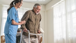 A nurse assists an elderly man with a walker in a bright room, highlighting hybrid care's role in modern healthcare.
