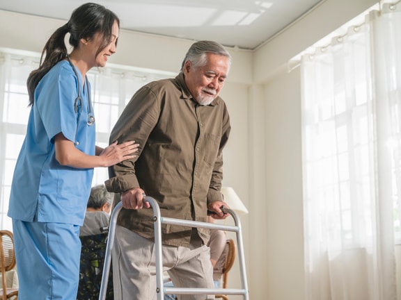 A nurse assists an elderly man with a walker in a bright room, highlighting hybrid care's role in modern healthcare.