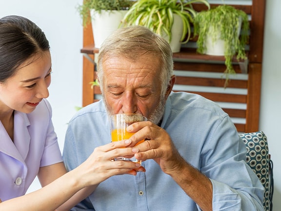 A medbridge caregiver helps an elderly man drink juice with potted plants in the background.