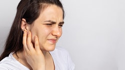 A woman in a white shirt looks pained while holding her ear, on a plain background.