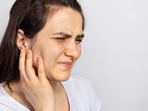 A woman in a white shirt looks pained while holding her ear, on a plain background.