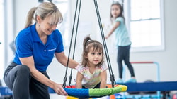 An adult assists a child on a swing in a Medbridge playroom, with another child on a platform nearby.