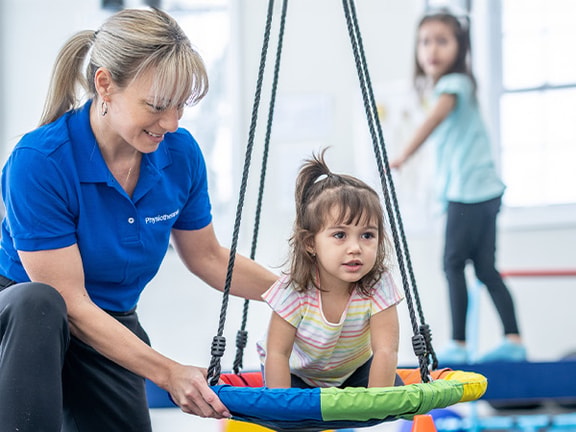 An adult assists a child on a swing in a Medbridge playroom, with another child on a platform nearby.