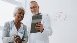 A doctor in a white coat presents a tablet to an elderly woman in a well-lit room with a Medbridge poster.