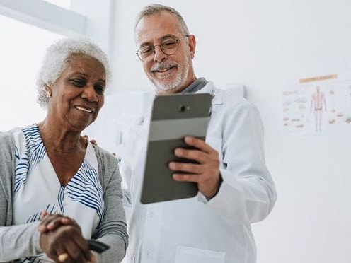 A doctor in a white coat presents a tablet to an elderly woman in a well-lit room with a Medbridge poster.