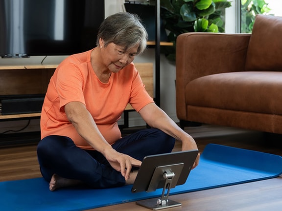 A person on a blue yoga mat in the living room uses a tablet with medbridge on it.