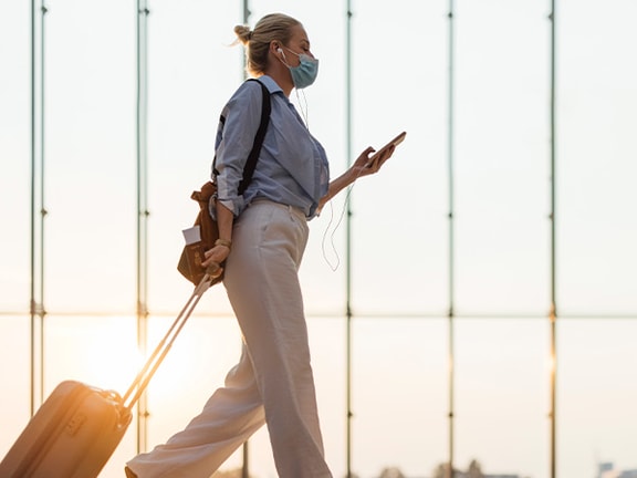 Traveler in face mask at airport, lit by sunlight through large windows.