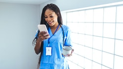 A healthcare professional in blue scrubs holds a smartphone and a mug near a glass block wall, representing medbridge.