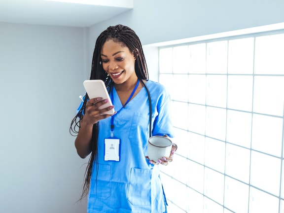 A healthcare professional in blue scrubs holds a smartphone and a mug near a glass block wall, representing medbridge.
