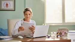 A doctor in a bright office reviews documents at a desk with a laptop, medical chart, and uterus model.
