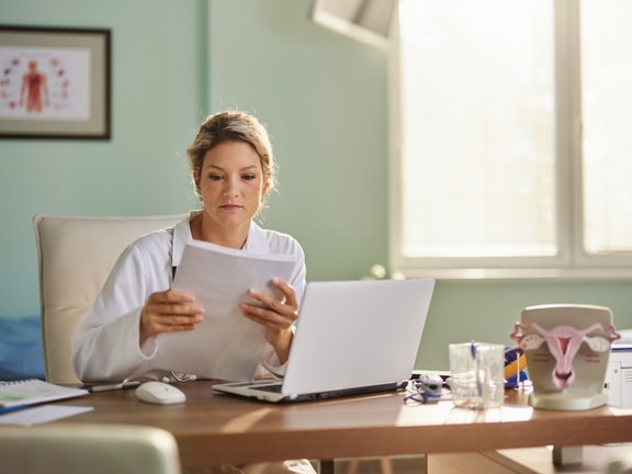A doctor in a bright office reviews documents at a desk with a laptop, medical chart, and uterus model.