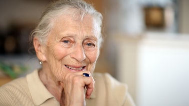 An elderly woman with gray hair and a beige shirt smiles gently indoors, reflecting the comfort medbridge hybrid care provides.