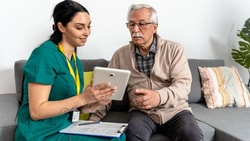 A clinician in green scrubs and an older man sit on a sofa looking at a tablet she holds, a clipboard on her lap.