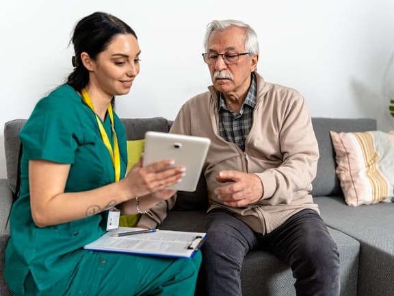 A clinician in green scrubs and an older man sit on a sofa looking at a tablet she holds, a clipboard on her lap.