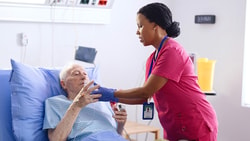 Speech-language pathologist in pink scrubs assisting an elderly patient in an ICU hospital bed, providing attentive care.