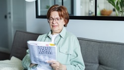 Older woman sitting on a gray couch, wearing glasses and solving a word puzzle to build cognitive reserve.