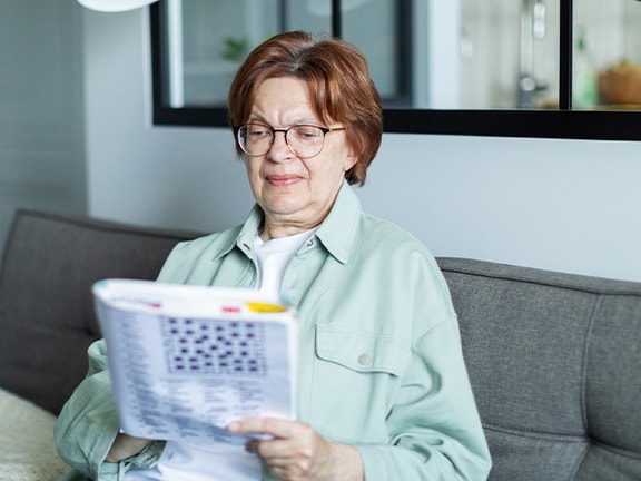 Older woman sitting on a gray couch, wearing glasses and solving a word puzzle to build cognitive reserve.