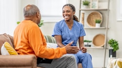 A smiling woman in scrubs chats with an older man on a couch in a well-lit room, part of medbridge's training.