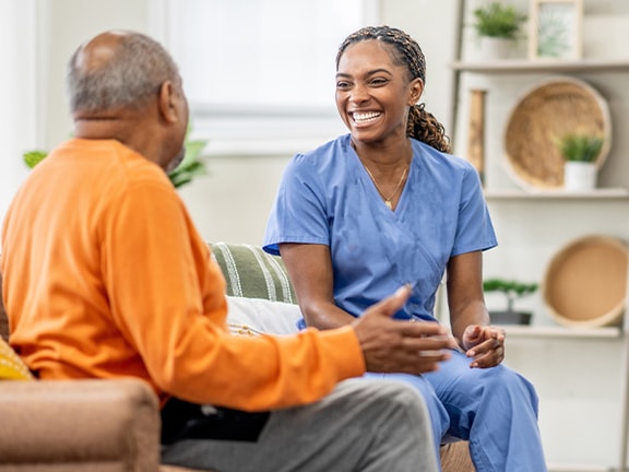 A smiling woman in scrubs chats with an older man on a couch in a well-lit room, part of medbridge's training.
