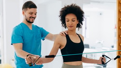 Physical therapist guiding a patient through resistance band shoulder exercises in a bright rehabilitation clinic.