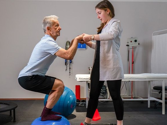 An older adult balances on two exercise balls with assistance at a Medbridge rehab center.