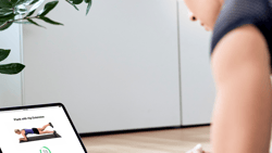 Person planking indoors while watching medbridge instructions on a laptop, with a large potted plant nearby.
