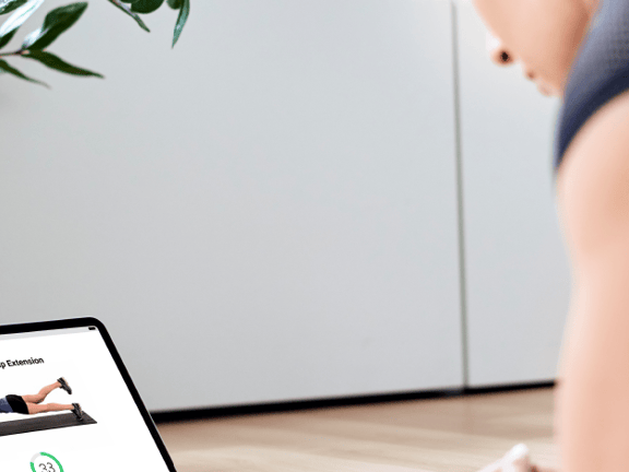 Person planking indoors while watching medbridge instructions on a laptop, with a large potted plant nearby.