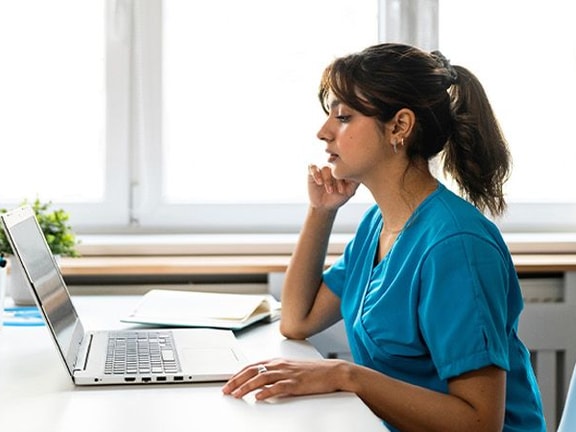 A woman in a blue shirt uses a laptop and talks on the phone in a bright room.