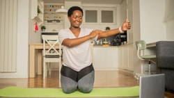 Person doing a chest stretch on a yoga mat while watching a laptop in a cozy apartment, with medbridge on screen.