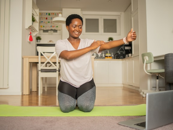 Person doing a chest stretch on a yoga mat while watching a laptop in a cozy apartment, with medbridge on screen.