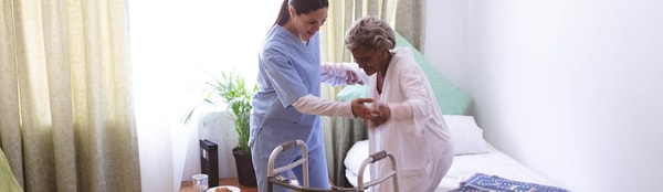 A caregiver helps an elderly woman with a walker in a bedroom.