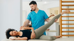 Physical therapist assisting a patient with a hip mobility exercise on a treatment table in a rehabilitation clinic.