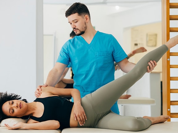 Physical therapist assisting a patient with a hip mobility exercise on a treatment table in a rehabilitation clinic.