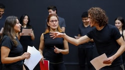 A group of people in casual black clothing stand in a studio discussing or rehearsing, holding papers, for medbridge.