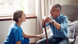 A Medbridge worker in blue scrubs talks to an elderly man with a cane on a couch.