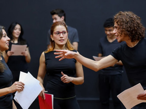 A group of people in casual black clothing stand in a studio discussing or rehearsing, holding papers, for medbridge.