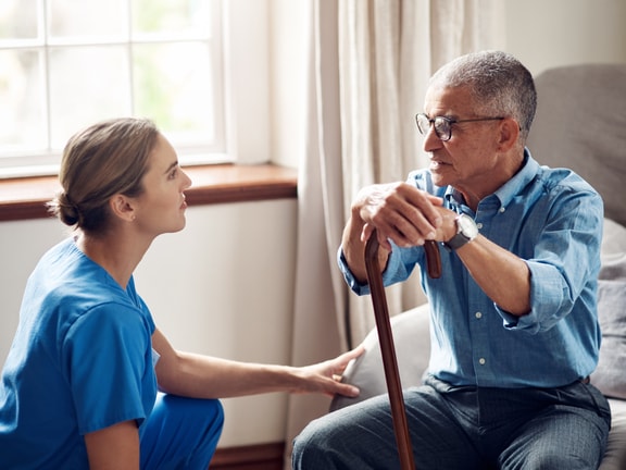 A Medbridge worker in blue scrubs talks to an elderly man with a cane on a couch.