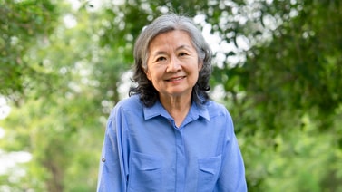 An older woman with gray hair and a blue shirt smiles outdoors with green trees, representing medbridge healthcare education.
