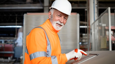 Older man in a hard hat and high-visibility jacket points to metal equipment, showing safety for medbridge healthcare training.