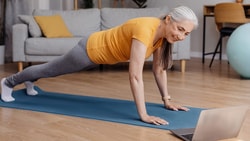 An older woman doing a plank on a yoga mat in her living room while following an online workout on her laptop.