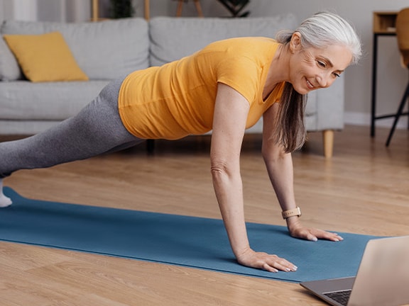 An older woman doing a plank on a yoga mat in her living room while following an online workout on her laptop.
