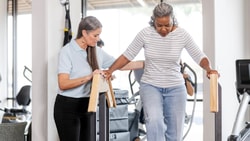 A woman helps another woman walk with parallel bars in a Medbridge rehab setting.